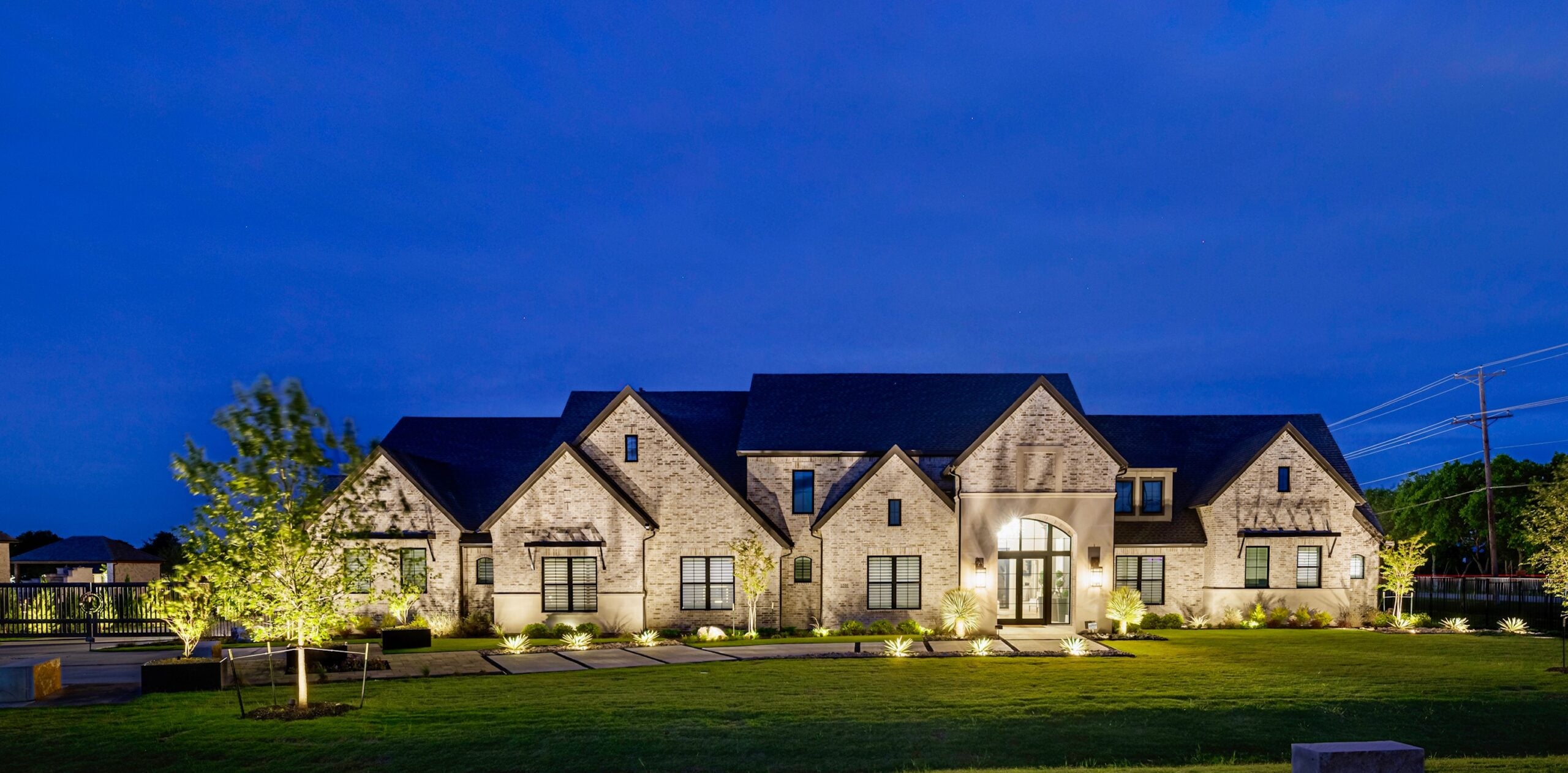 A modern brick house with multiple gables, illuminated landscaping, and a twilight sky in the background.