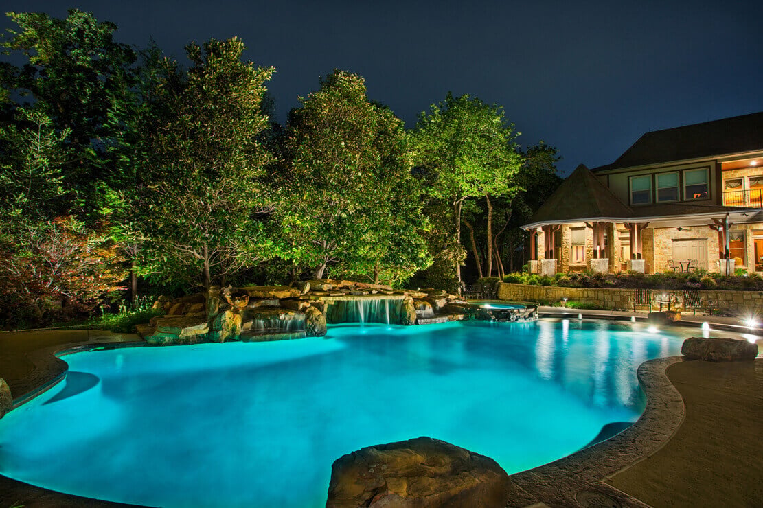 A resort-style swimming pool featuring bright blue water lighting framed by dense uplit trees along the property edge.