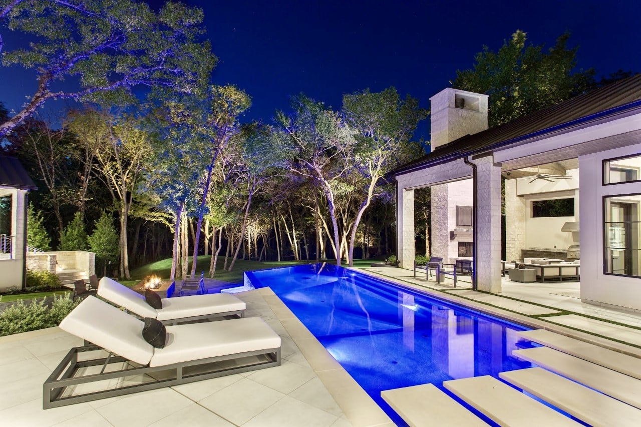 Modern backyard with a brightly lit pool, loungers, and lush trees under a night sky.