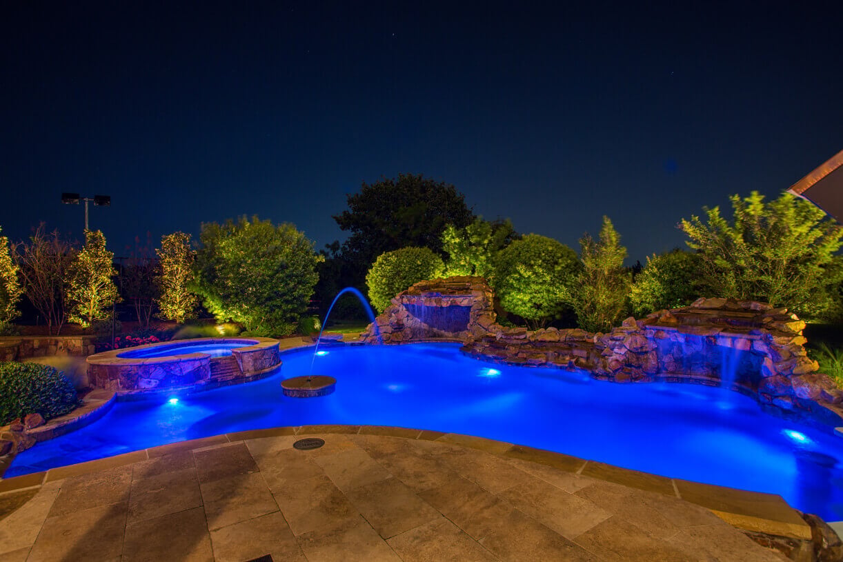 A custom backyard pool with blue underwater LED lighting, stone rock waterfalls, and warm landscape tree accents.