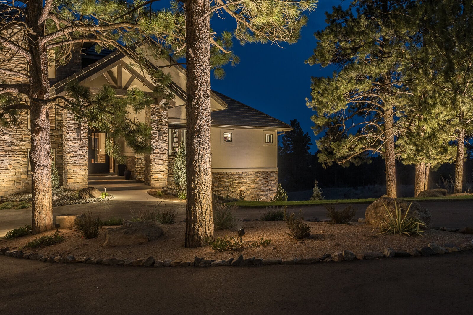 A modern house at night, illuminated by lights, surrounded by tall trees and landscaped gardens.