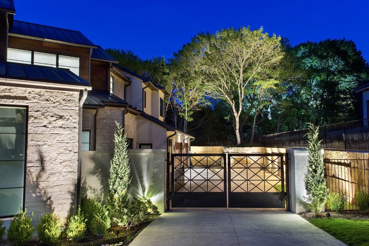 Night view of a modern home entrance with illuminated gate, surrounded by trees and landscaped shrubs.