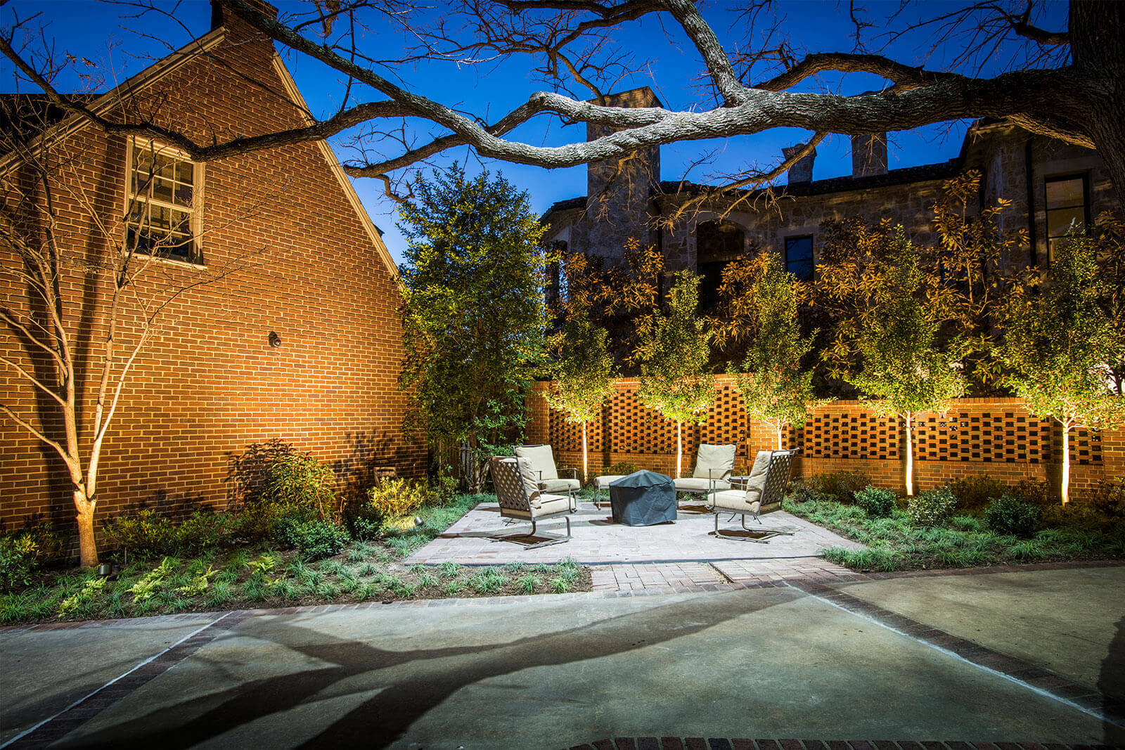 An outdoor patio with seating and a central fire pit set against a brick wall, surrounded by festive uplit trees and warm holiday ambient lighting.