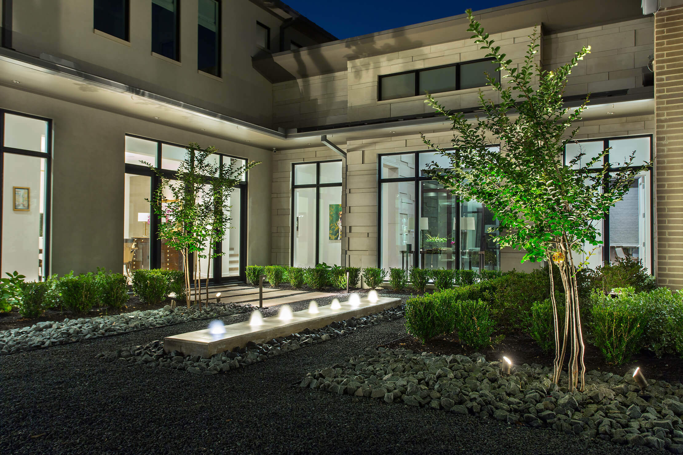 Modern courtyard at night featuring plants, stone pathways, and illuminated lighting elements.
