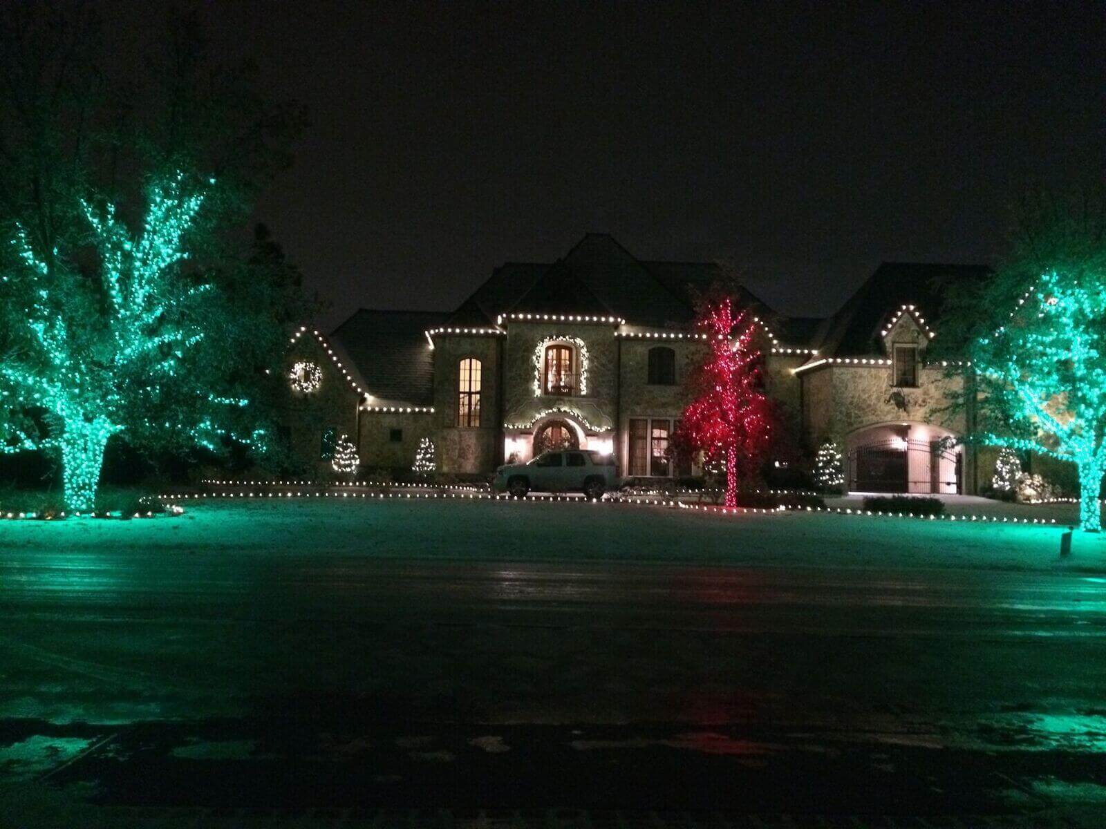 A large, illuminated house with green and red holiday lights on trees and decorations in the yard at night.