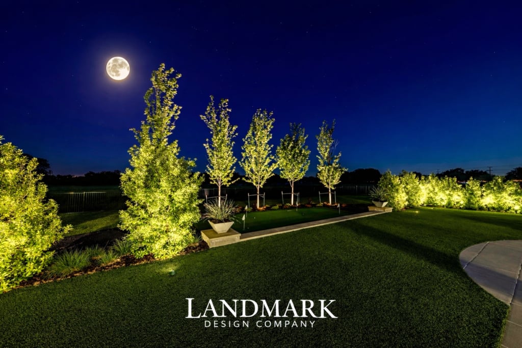 A night sky with a full moon above a well-lit landscaped garden featuring trees and green grass.