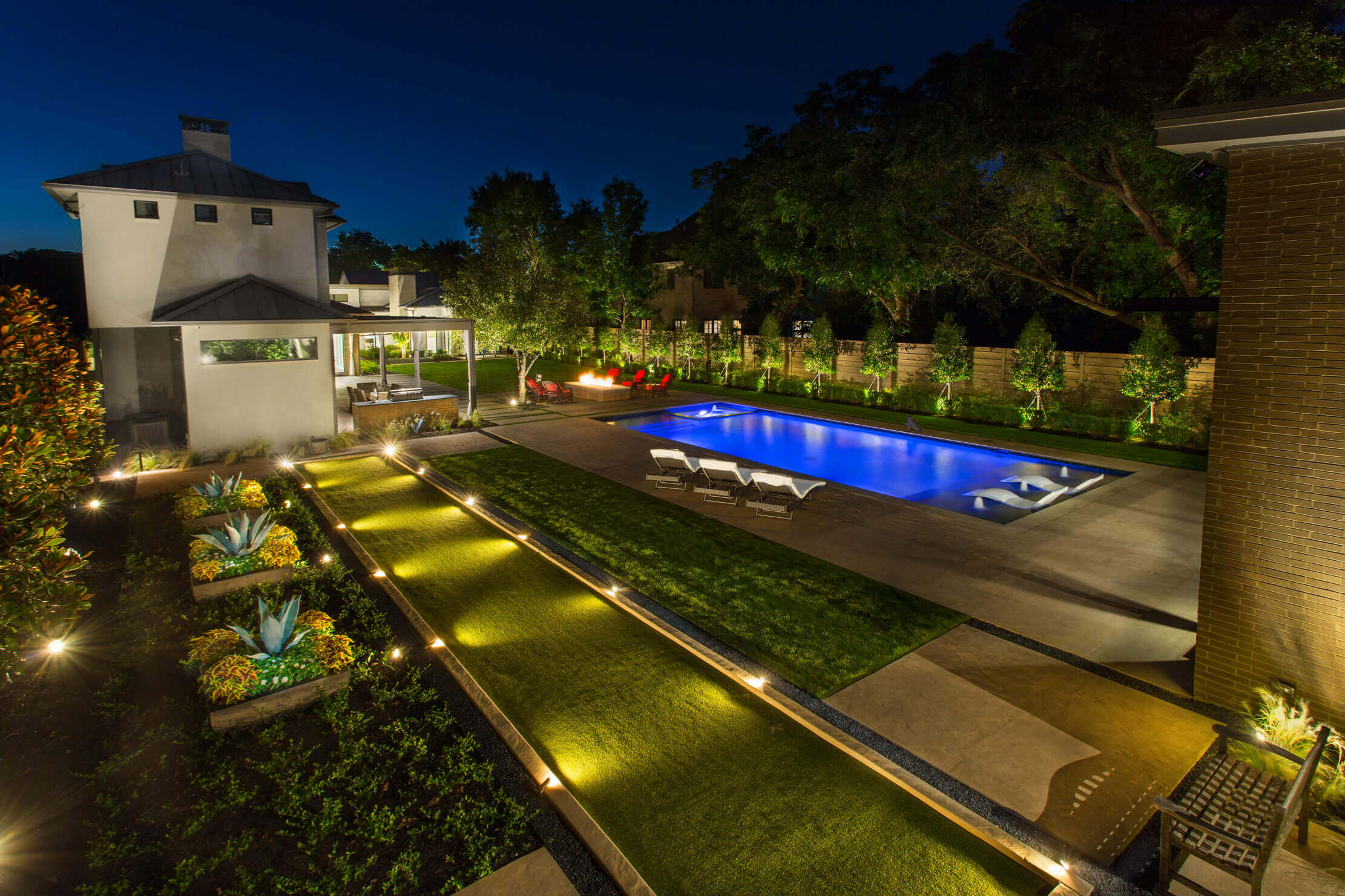 A tranquil backyard at night featuring a lit pool, landscaped garden, and modern house with outdoor seating.