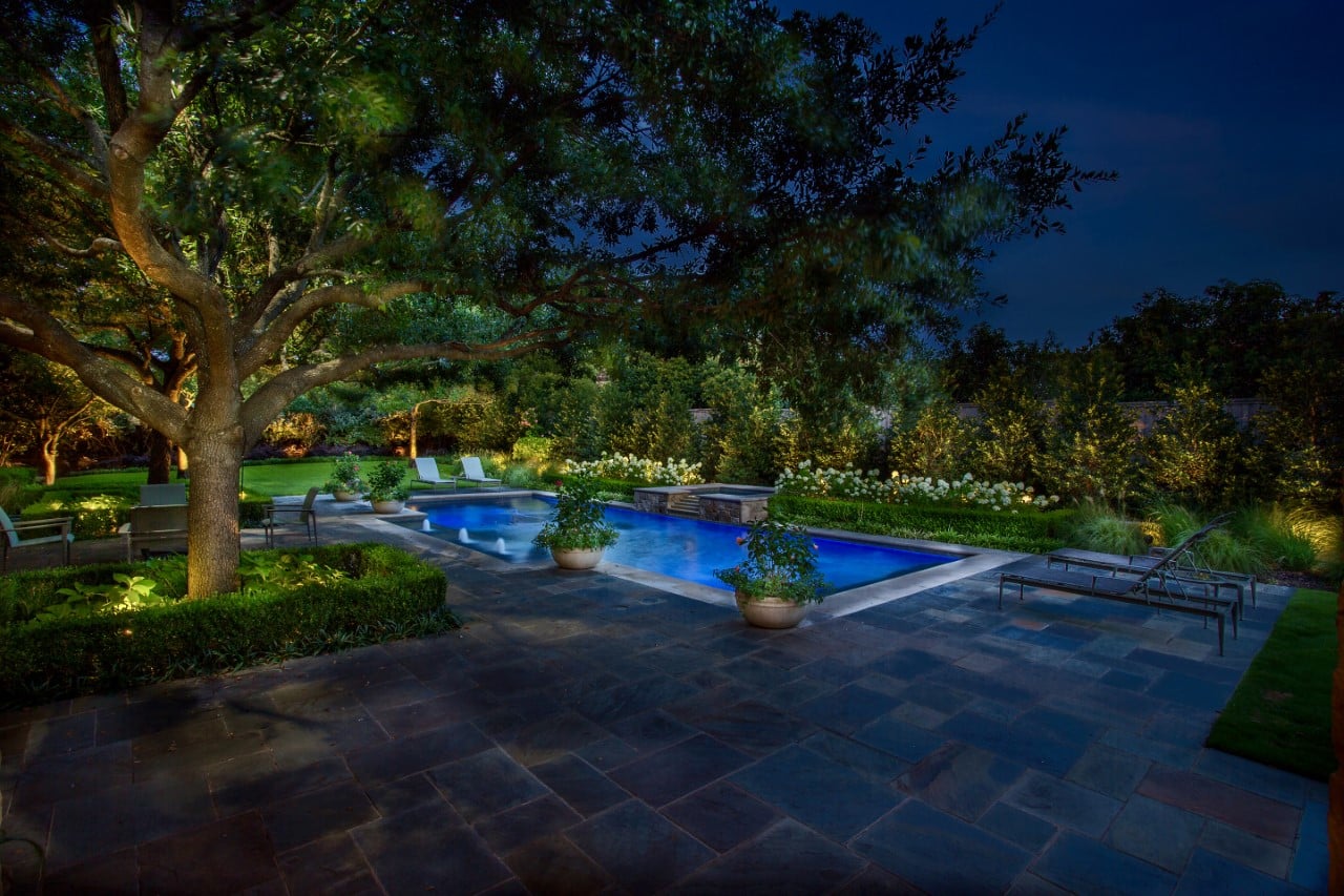 Peaceful backyard at night featuring a swimming pool, surrounded by trees and illuminated plants.
