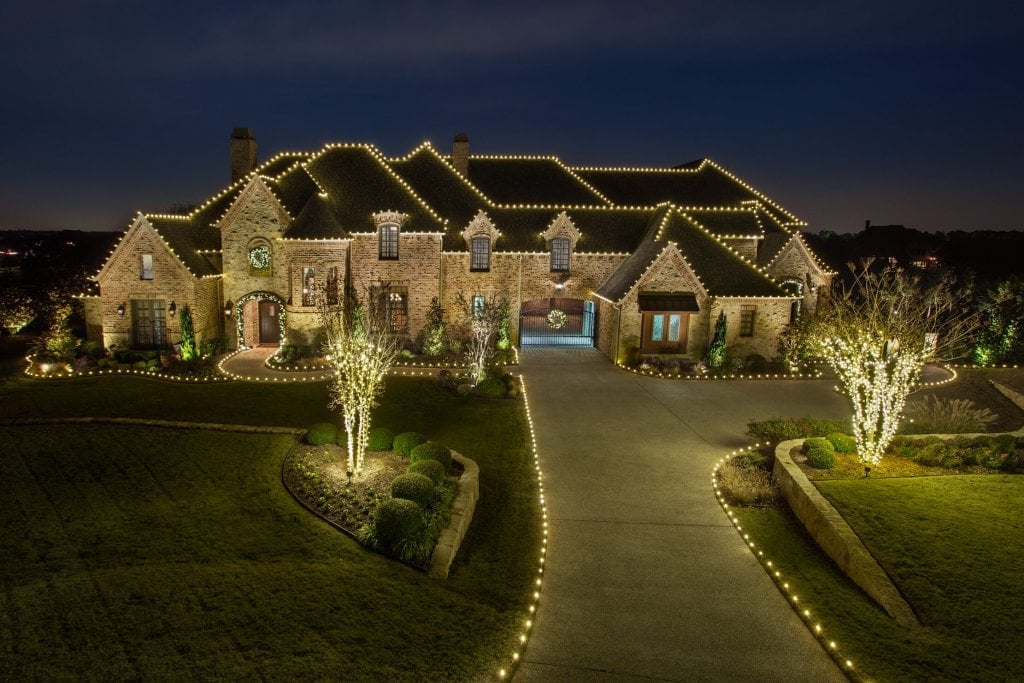 A large stone house decorated with string lights, surrounded by landscaped lawns and illuminated trees at night.