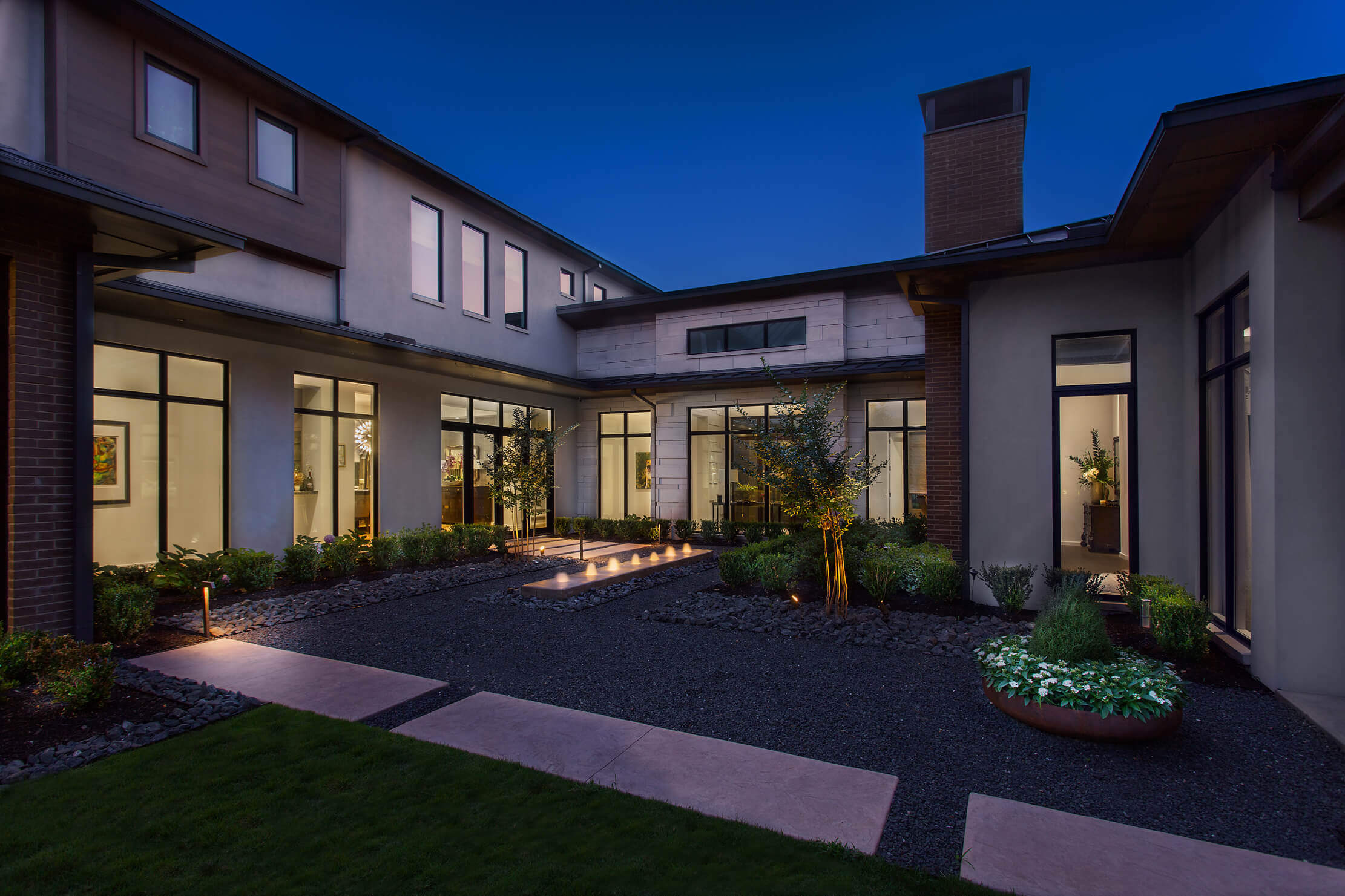 A modern courtyard with landscaped gardens and pathways, illuminated at dusk, surrounded by building facades.