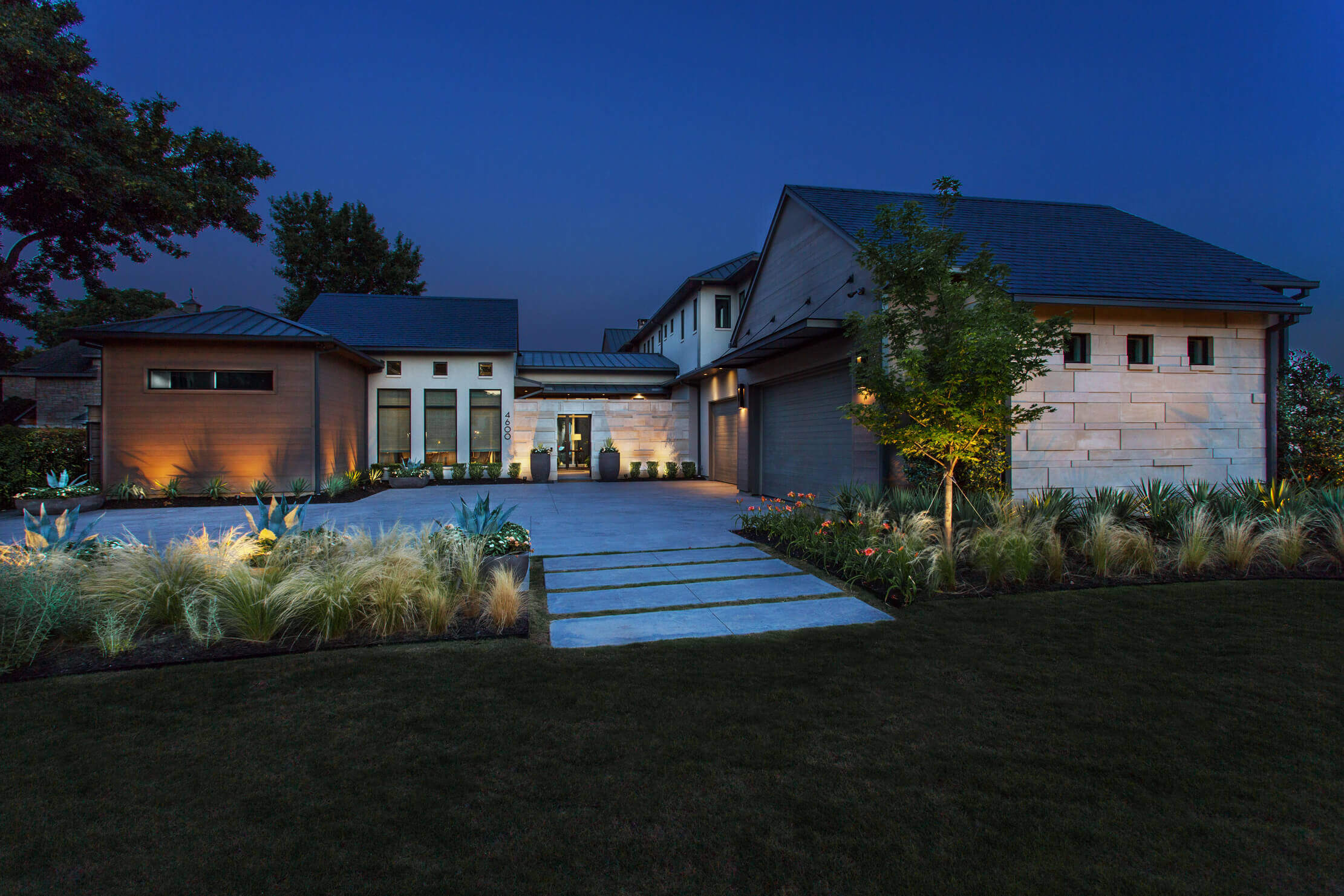 Modern house exterior at dusk, featuring stone and wood materials, with landscaped garden and pathway.