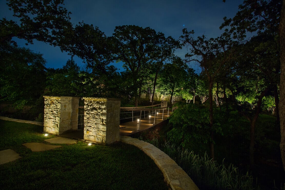 Stone entryway pillars and a walkway illuminated with ambient landscape lighting framed by dark surrounding trees.