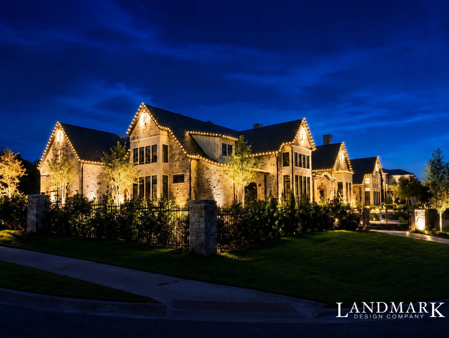 Stone home across a wide lawn with warm white lights tracing the full roofline and a lit driveway in front.