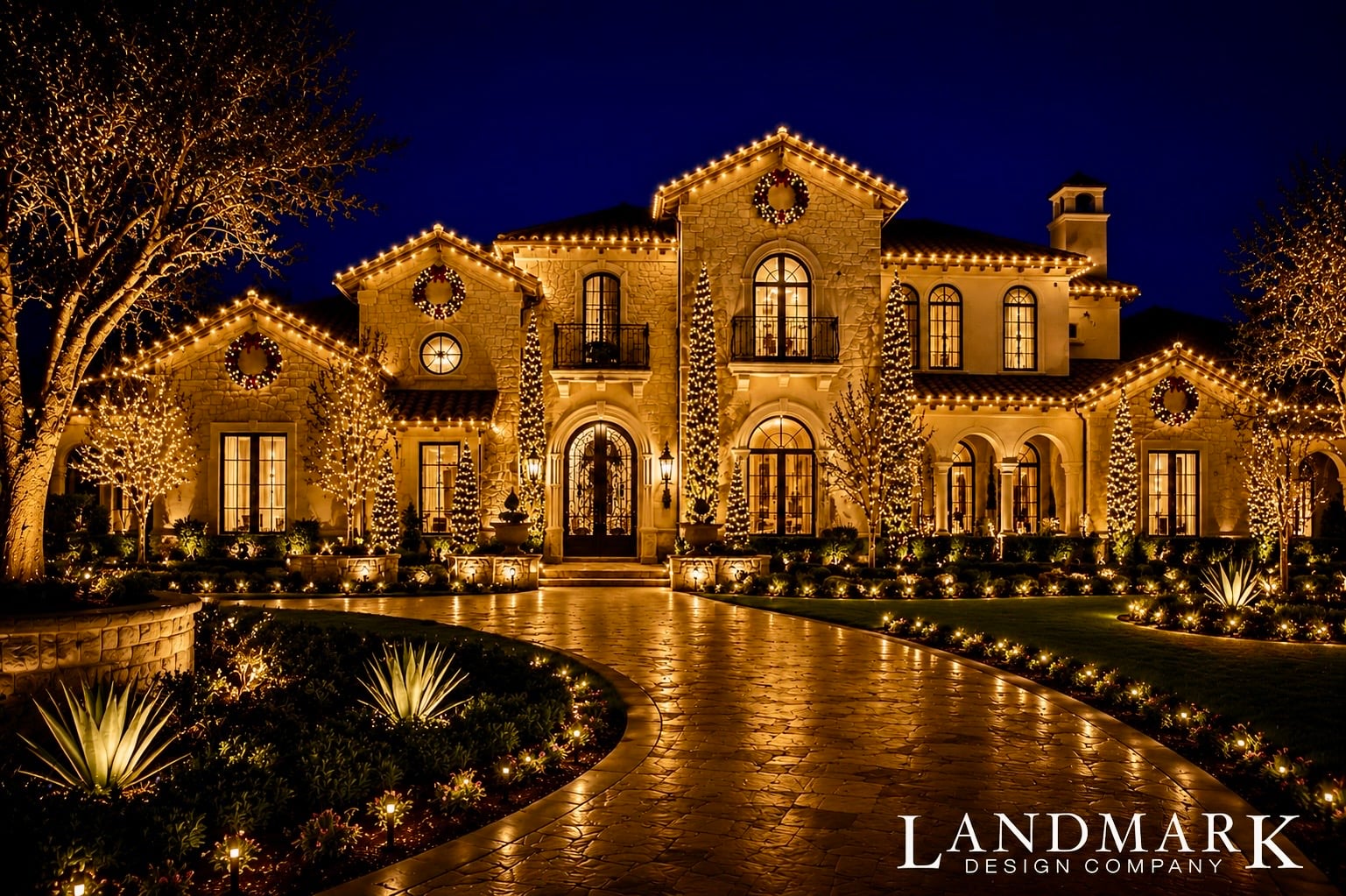 A beautifully lit luxury home at night, decorated with holiday lights and surrounded by landscaped greenery.