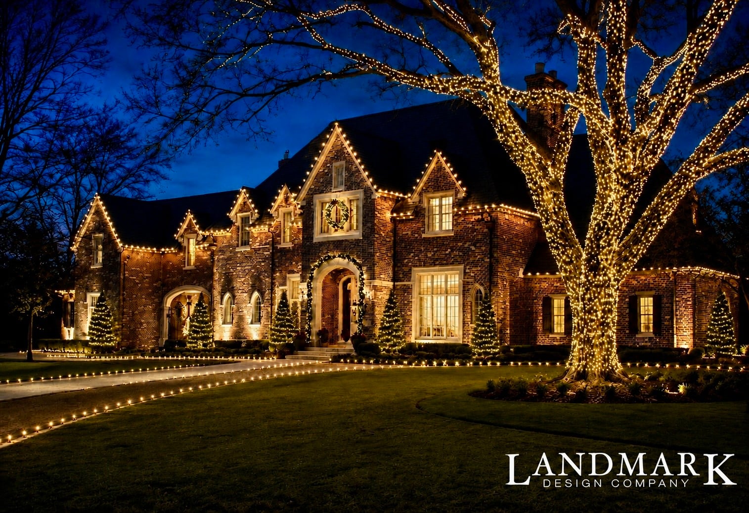 A beautifully lit brick house at night with festive lights and small trees. A well-maintained lawn is visible.