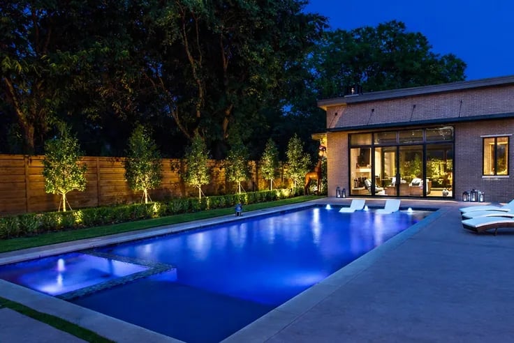 Lit pool and modern pool house at dusk, with uplit trees along a wood privacy fence