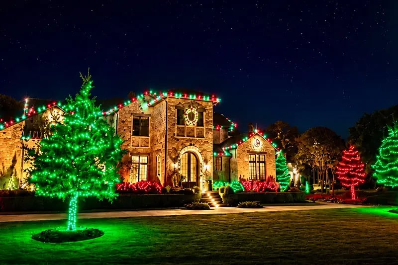 Stone home with roofline Christmas lights and wreaths, a green-wrapped tree and red-lit trees on the lawn