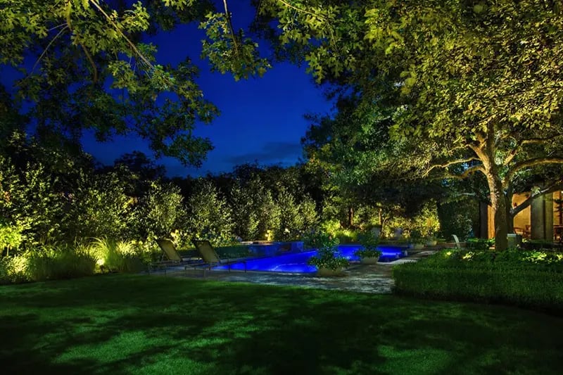 Uplit oak canopies over a lawn and planted beds at dusk, with a glowing pool beyond