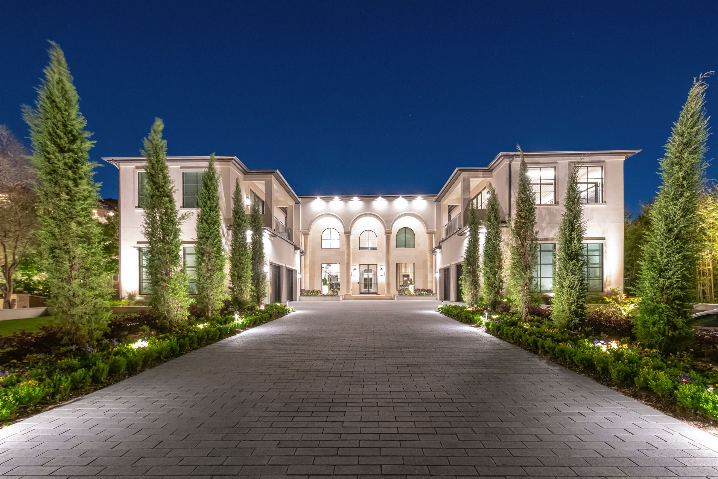 Two-story estate at night with warm architectural lighting on the facade and uplit cypress trees along the drive