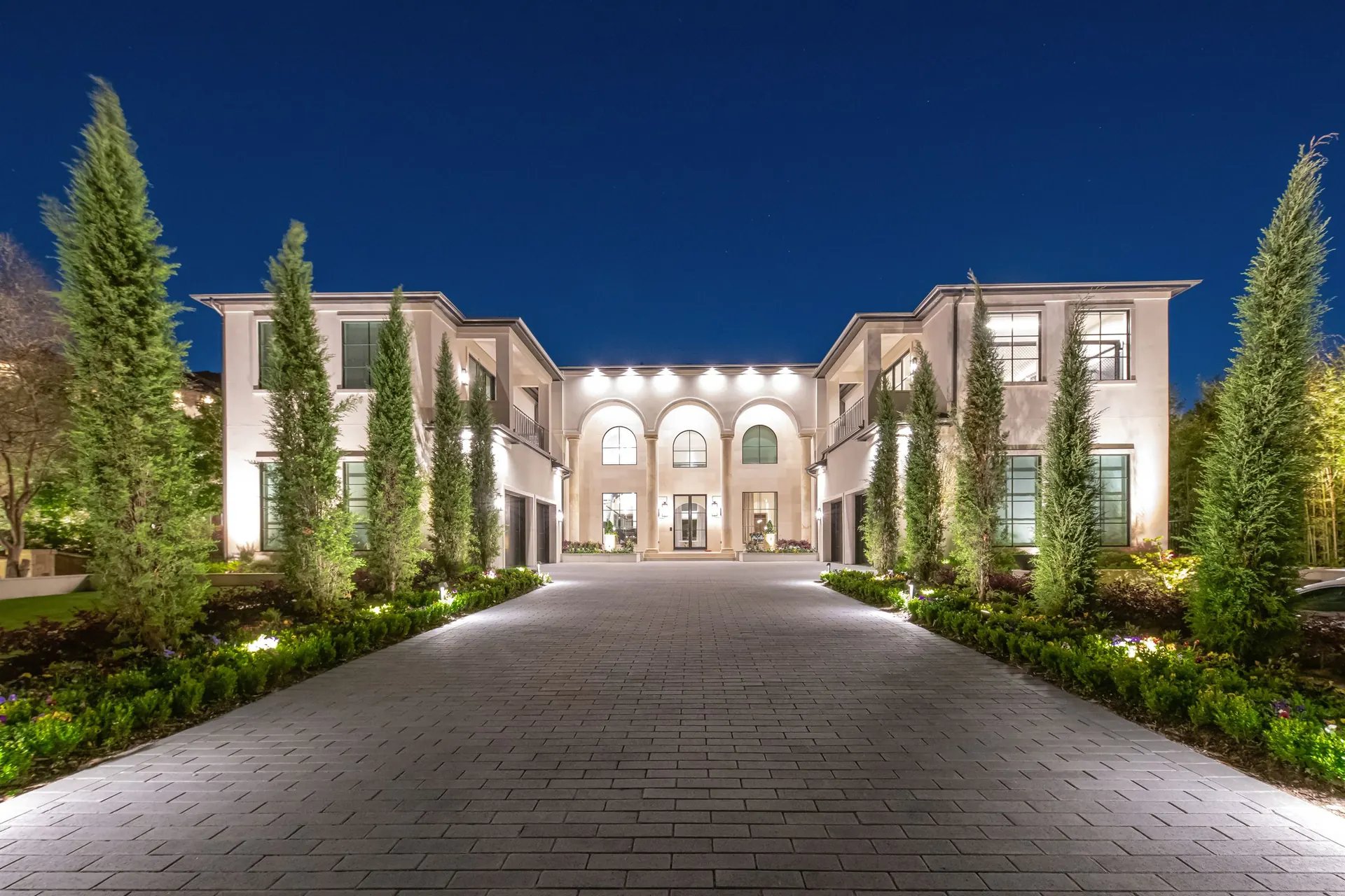Two-story estate at night with warm architectural lighting on the facade and uplit cypress trees along the drive
