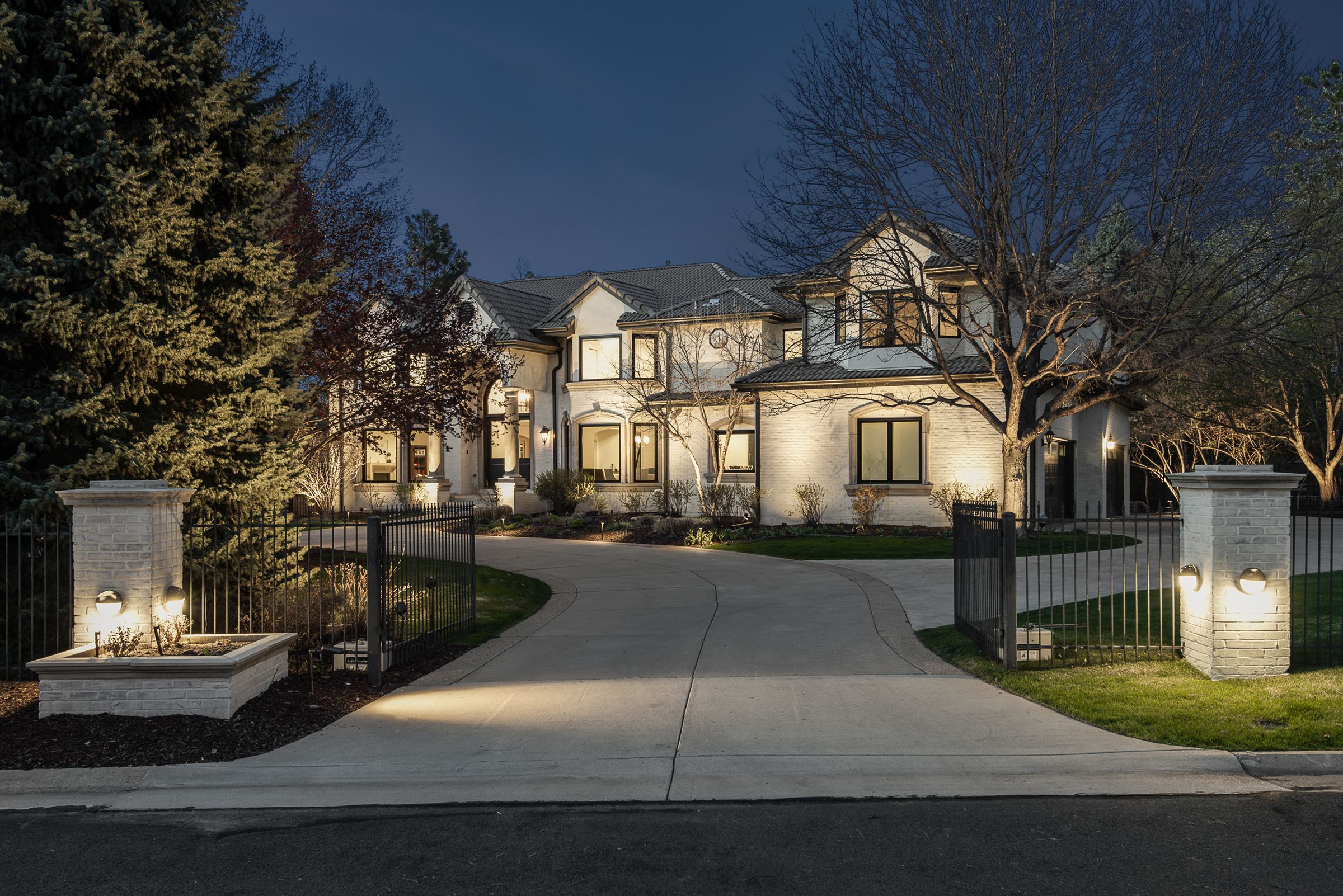 White two-story home at dusk with lit pillars along a gated circular drive