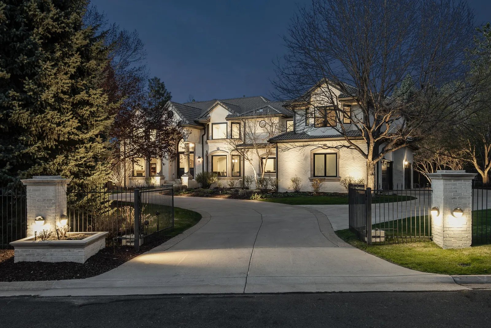 White two-story home at dusk with lit pillars along a gated circular drive