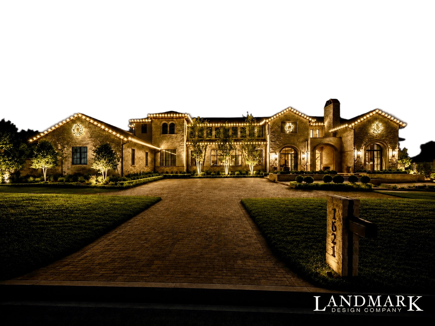 Stone Mediterranean-style home outlined in warm roofline lights, with uplit trees along a paver driveway at night
