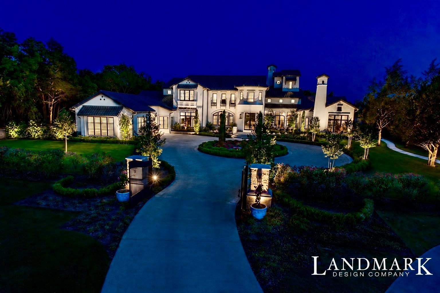 White two-story home at night with a lit circular drive and uplit trees